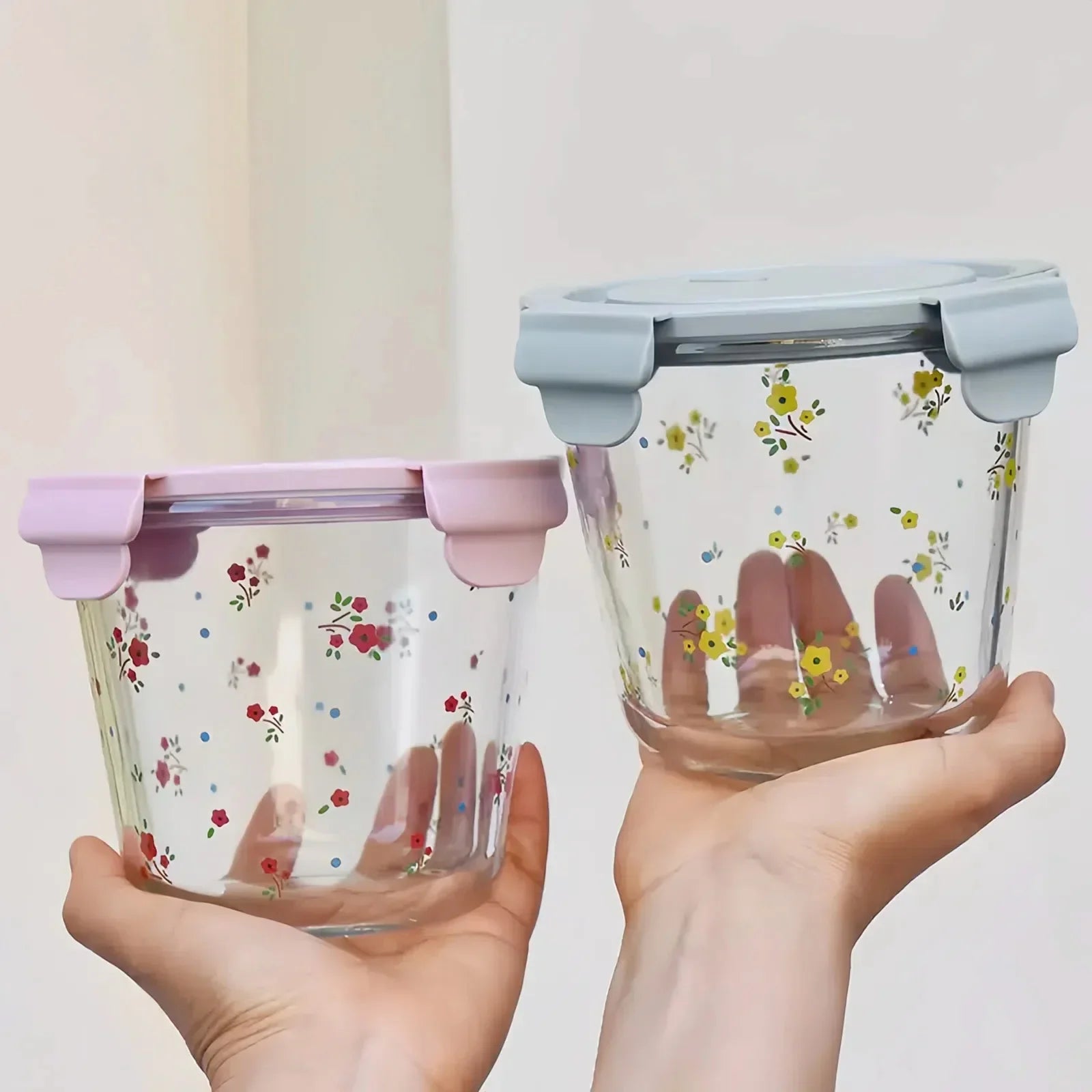Two clear glass food storage containers with floral patterns and pastel lids held in hands