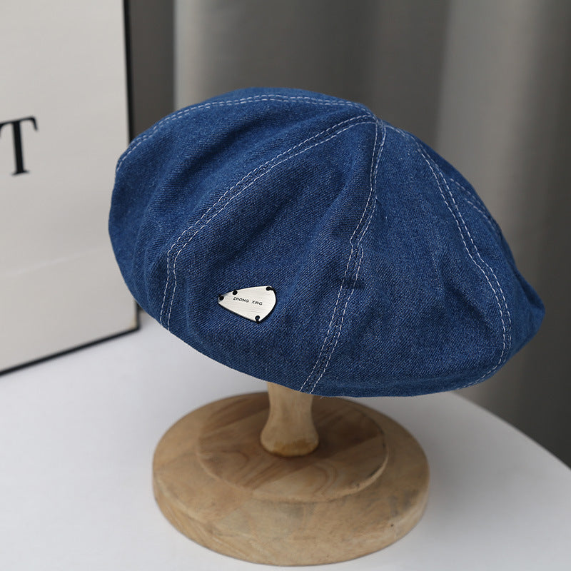 Denim beret hat with white stitching and metal accent on a wooden display stand