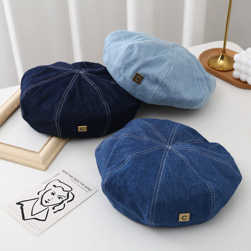 Three denim berets in light, medium, and dark blue with metal tags on a white table.