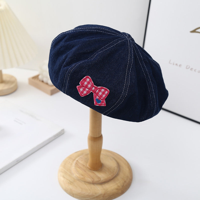 Denim beret hat with red checkered bow patch on display stand in minimalist room