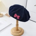 Denim beret hat with red checkered bow patch on display stand in minimalist room