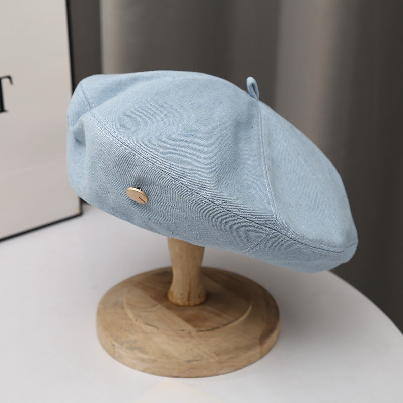 Light blue denim beret hat with metal button accent on a wooden stand, minimalist style