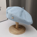 Light blue denim beret hat with metal button accent on a wooden stand, minimalist style