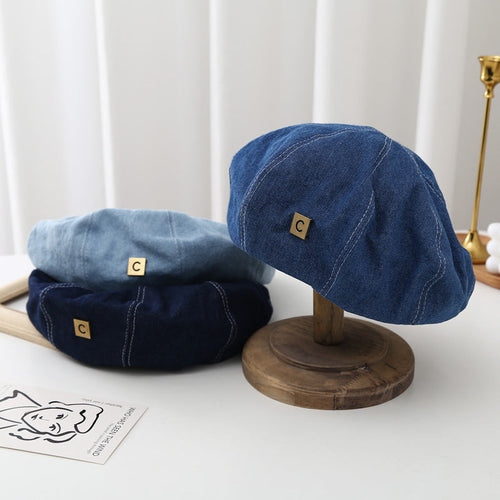 Set of three denim berets in light, medium, and dark blue with gold C label, on display stand
